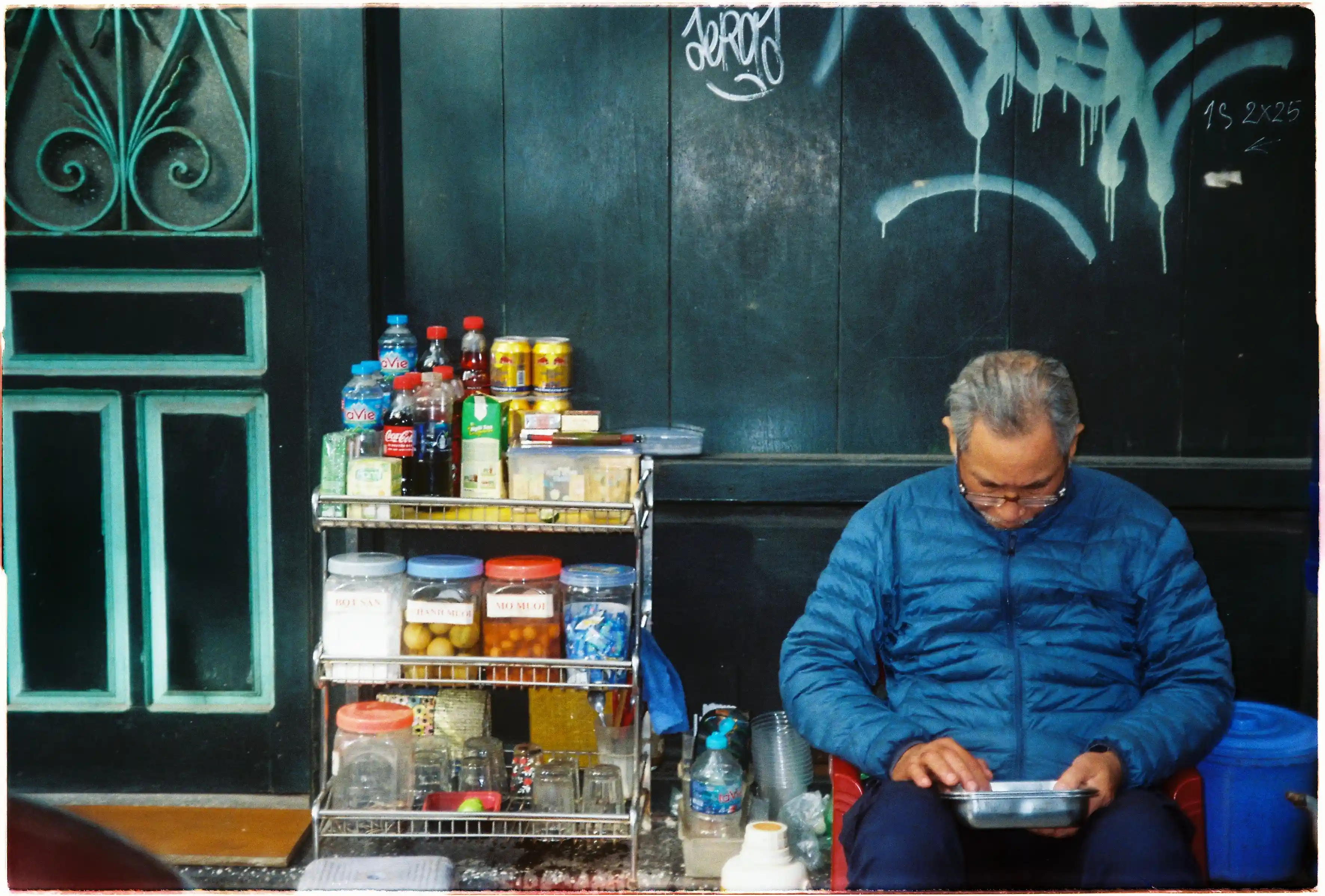 Cụ ông mặc áo phao xanh bên quầy nước vỉa hè: Ảnh chân dung đường phố chụp tại tiêu cự 60mm của Canon Autoboy Tele 6