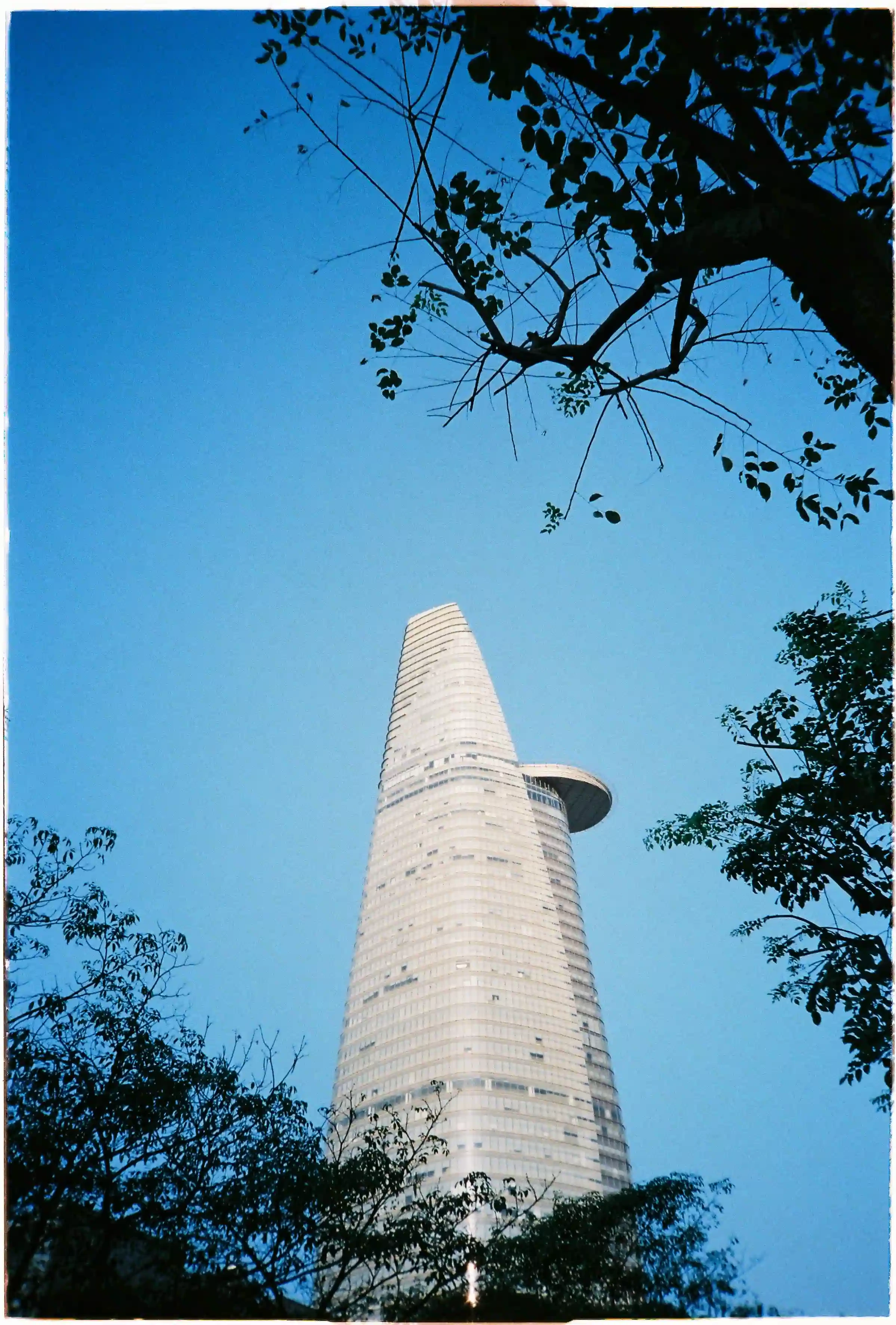 Tháp Bitexco nổi bật trên nền trời xanh ngắt và tán lá xanh rì: Ảnh phong cảnh xuôi sáng rực rỡ chụp bởi Fuji Cardia Everyday OP