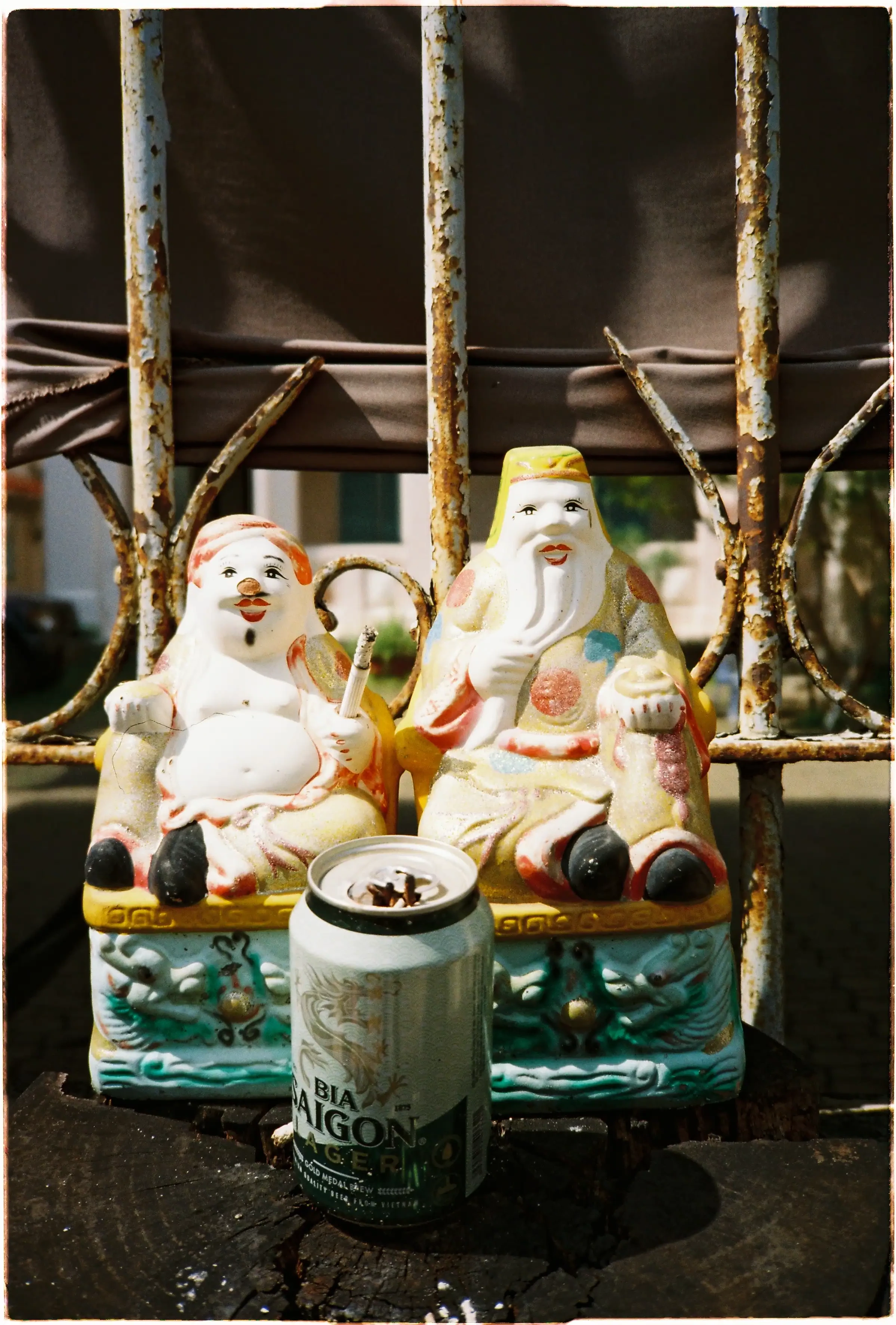 Tượng Ông Địa và Thần Tài phơi nắng: Ảnh chụp xuôi sáng sắc nét, tương phản cao từ máy film Canon Autoboy 3 - Kodak Gold 200