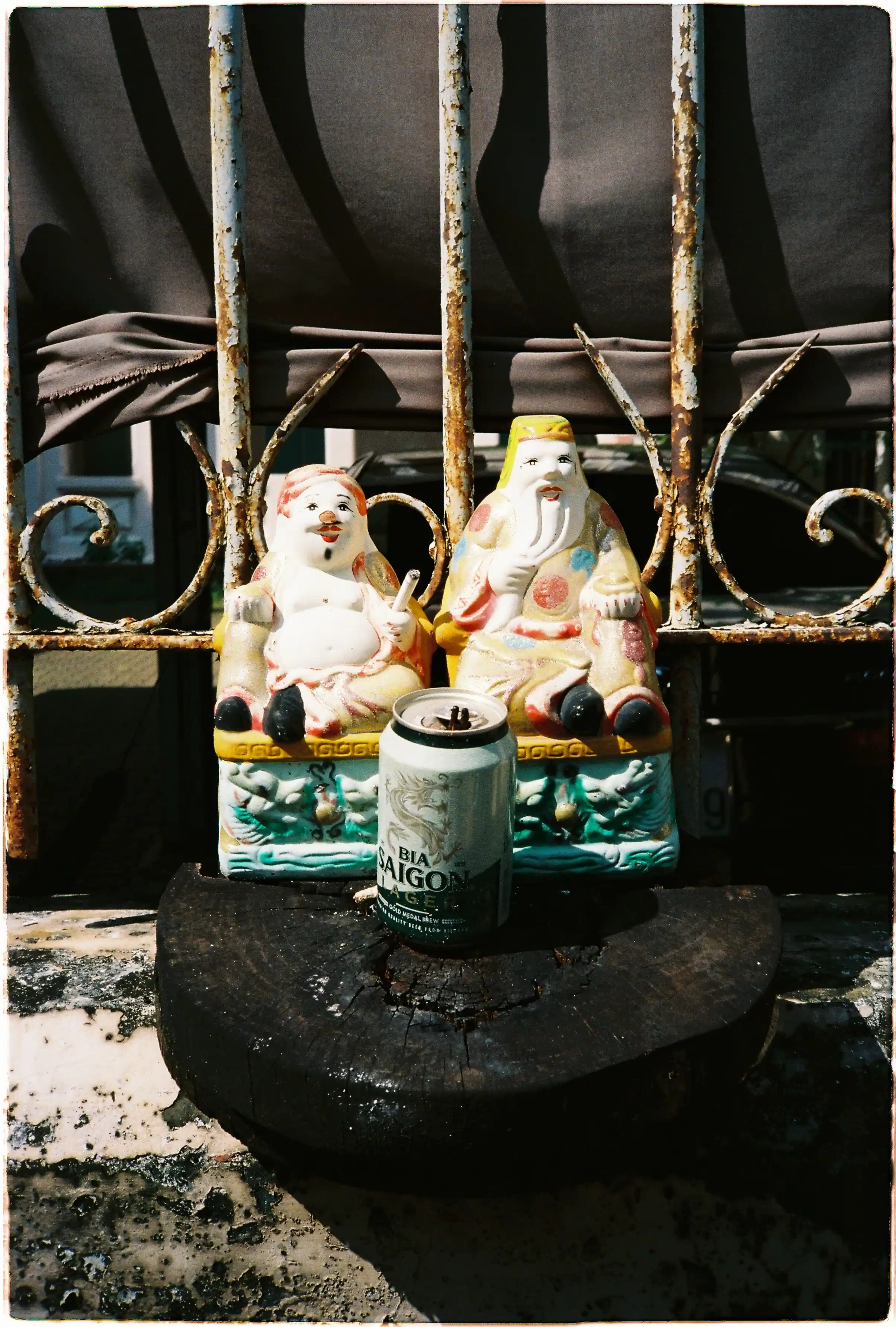 Ảnh tĩnh vật hài hước: Tượng Ông Địa Thần Tài và lon bia Sài Gòn, ảnh chụp sắc nét làm nổi bật chất liệu sứ và gỉ sét từ Nikon L35AD3 - Fuji 400