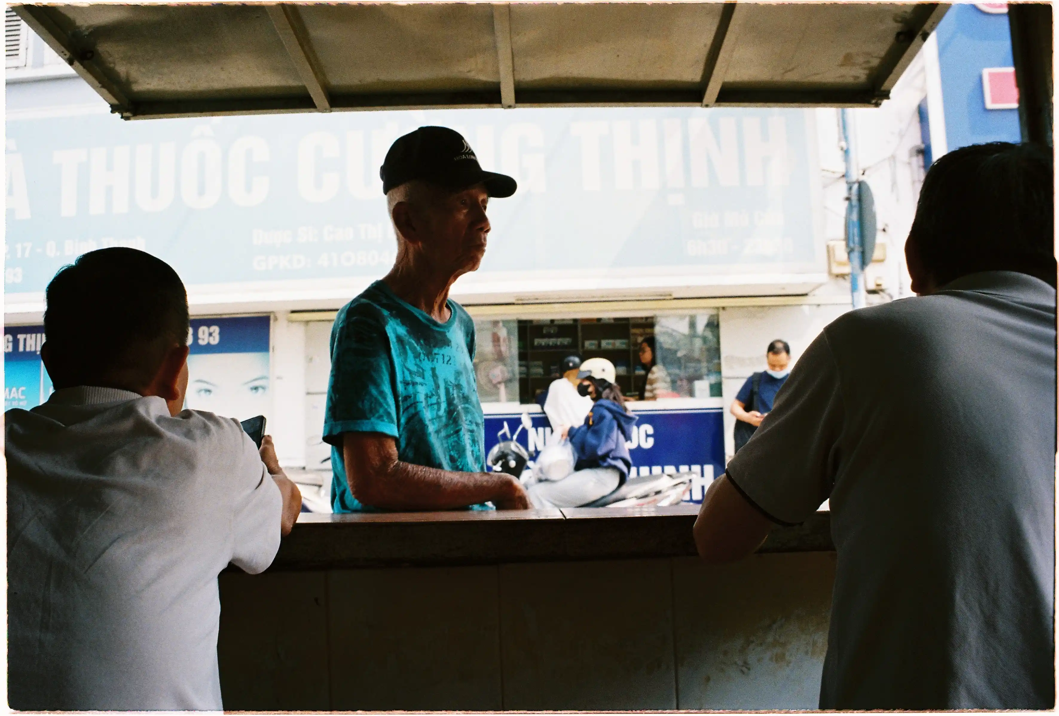 Cụ ông mặc áo xanh đứng giữa hai người ở tiền cảnh: Bố cục đóng khung (framing) tự nhiên trong ảnh đời thường chụp bởi Canon Autoboy Jet