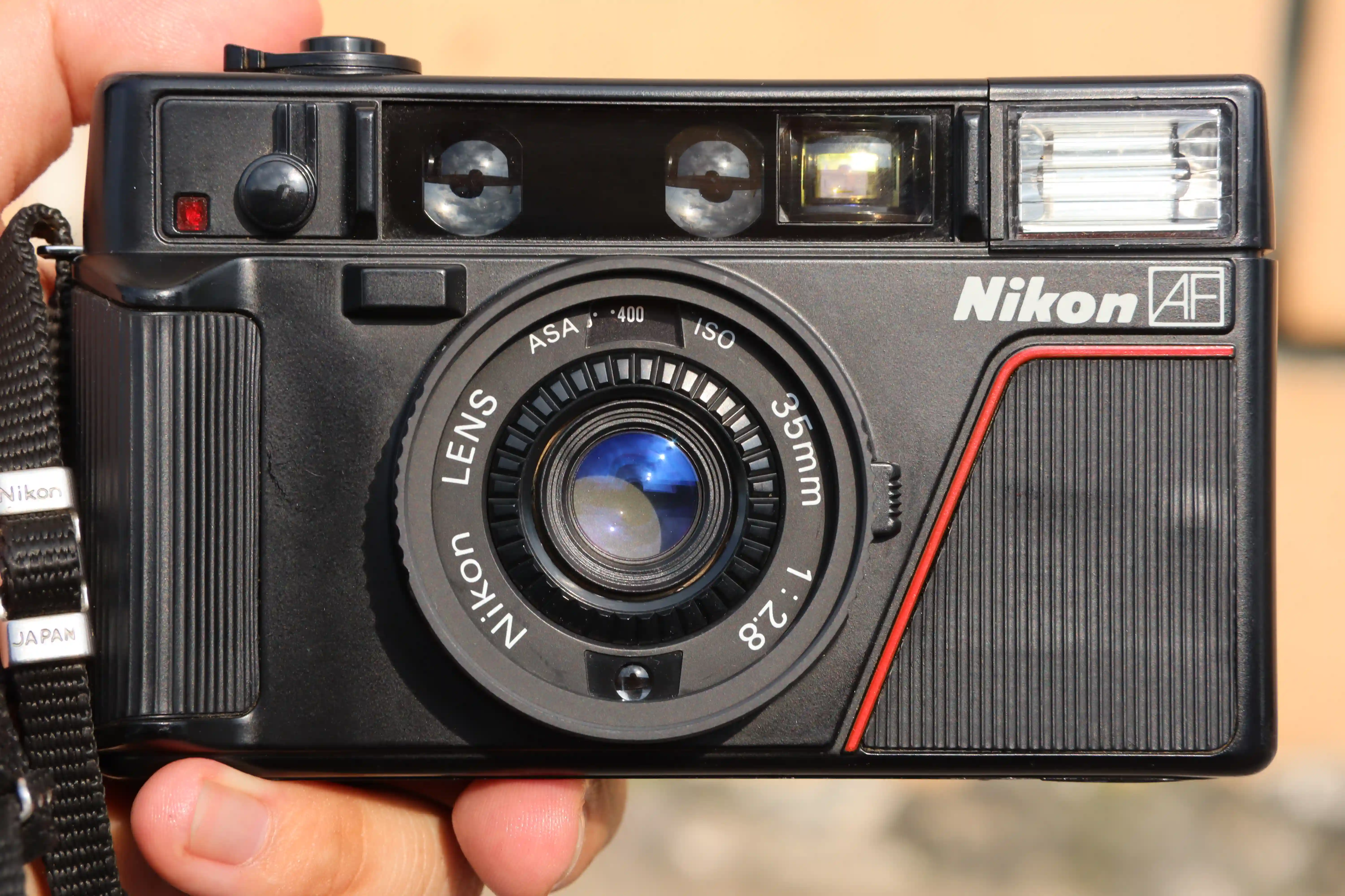 Mặt trước của máy ảnh film cổ điển Nikon L35AF, với ống kính, đèn flash, logo Nikon AF, vòng xoay chỉnh iso, cần gạt hẹn giờ và mắt đo sáng ở dưới ống kính.